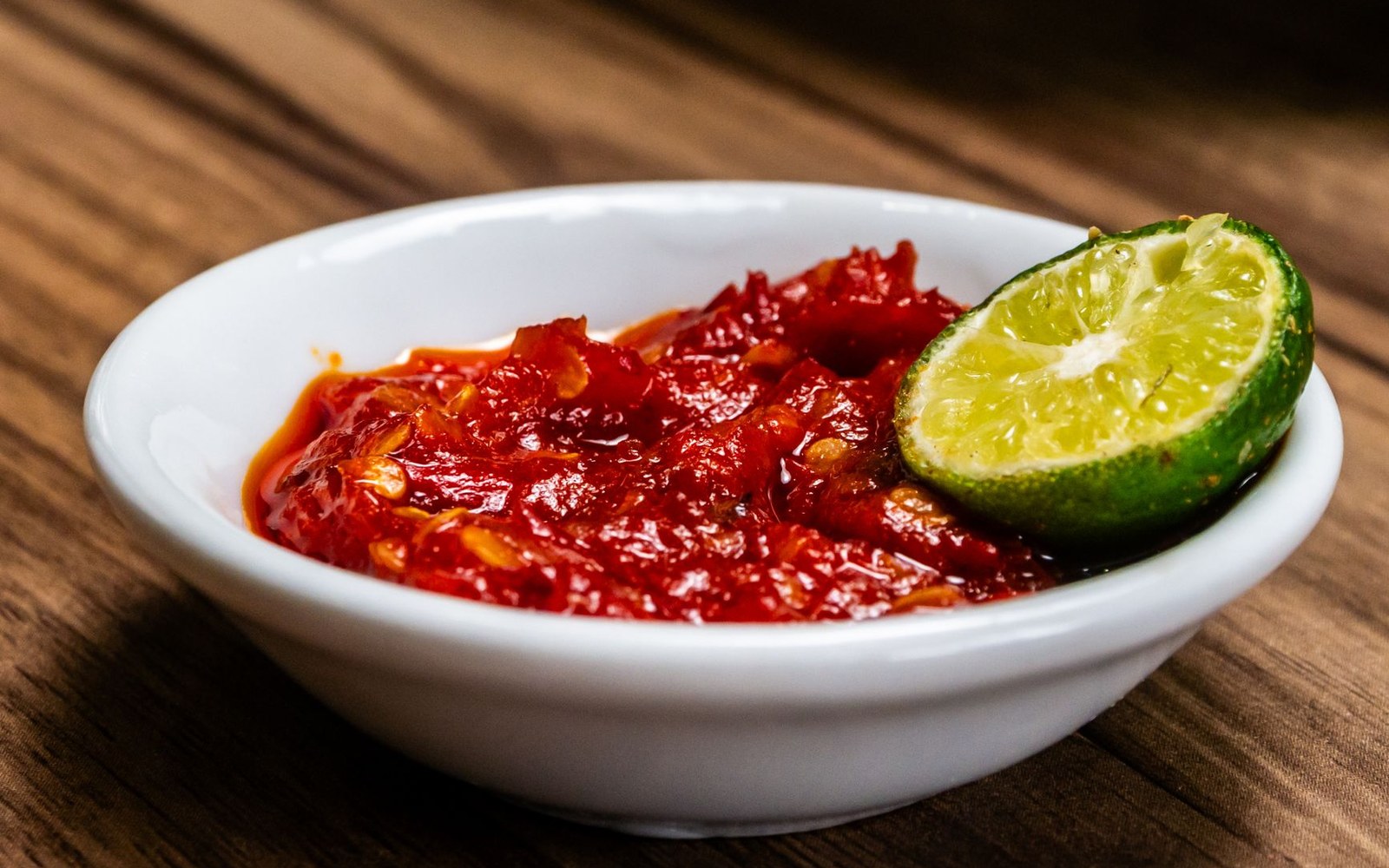 White bowl of red chile sauce topped with a halved lime on a wooden table