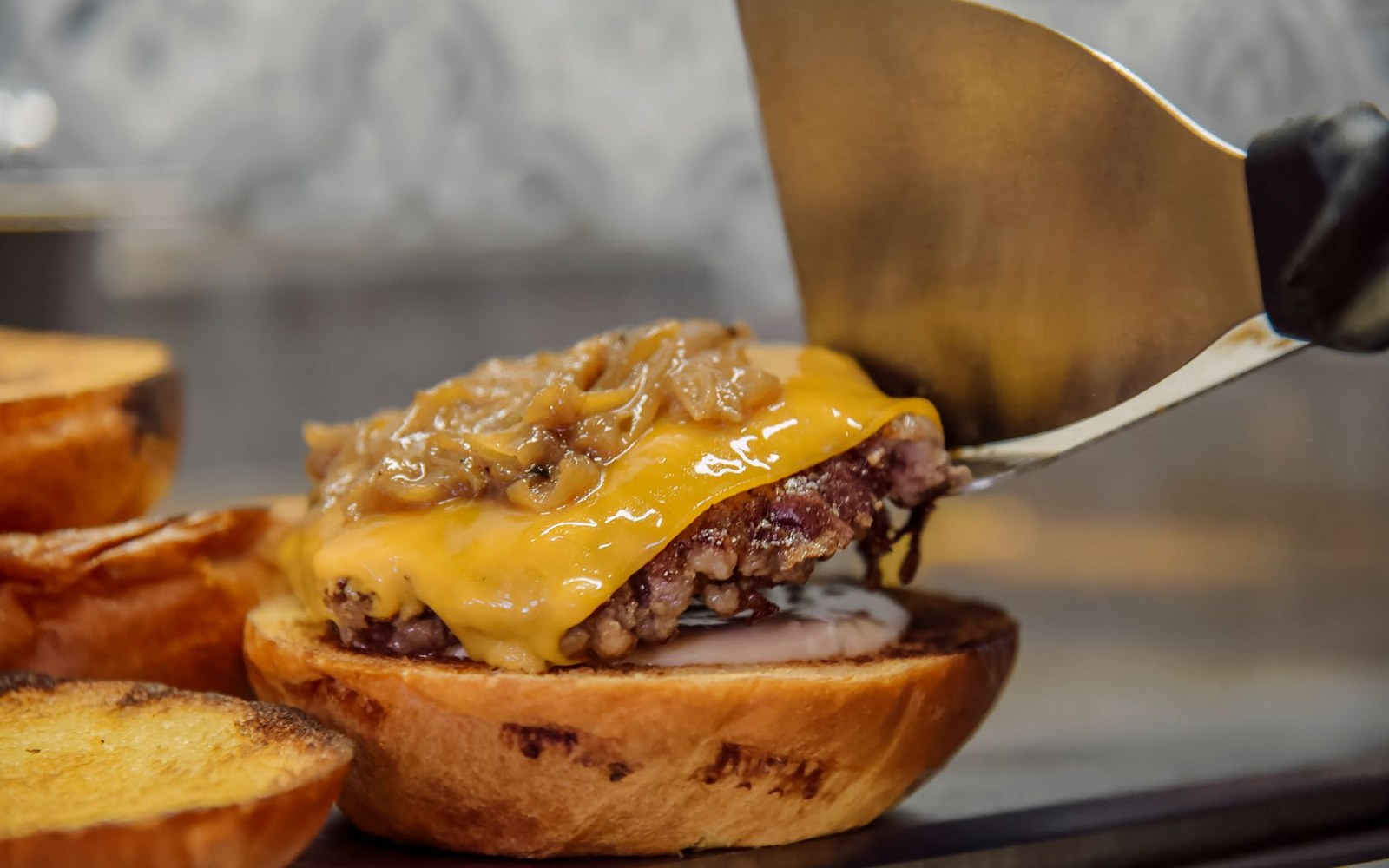 A spatula lifts a cheeseburger patty topped with melted cheddar and sautéed onions onto a toasted bun