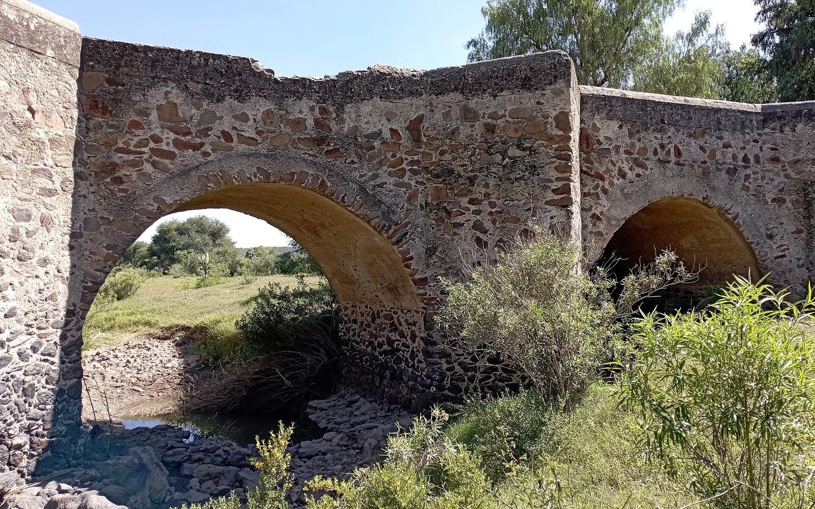 Old stone bridge with two arches spanning a shallow creek surrounded by brush