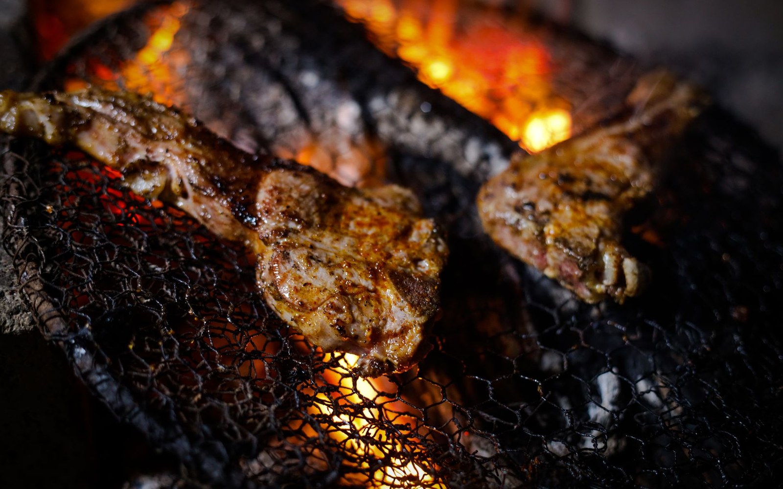 Meat cuts sear on a wire grill over glowing embers and open flame